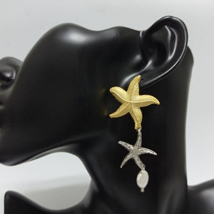 May include: A drop earring featuring a textured gold-tone starfish at the top, connected to a smaller silver-tone starfish below, which is accented with a single white oval-shaped pearl dangling from the bottom of the silver starfish.