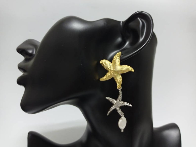 May include: A drop earring featuring a textured gold-tone starfish at the top, connected to a smaller silver-tone starfish below, which is accented with a single white oval-shaped pearl dangling from the bottom of the silver starfish.