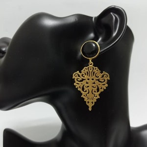 May include: Gold-colored filigree earrings with a black circular stud. The earrings feature an ornate, symmetrical design. The earrings are displayed on a black mannequin head against a white background.