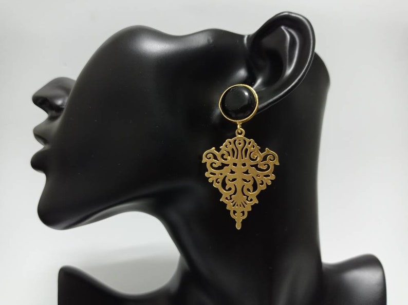 May include: Gold-colored filigree earrings with a black circular stud. The earrings feature an ornate, symmetrical design. The earrings are displayed on a black mannequin head against a white background.