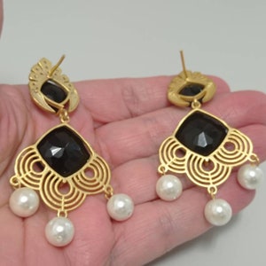 May include: Gold-coloured chandelier earrings featuring a black, faceted central stone and three dangling white pearls. The earrings have an openwork design and a post backing. The earrings are held in a hand.
