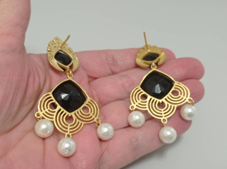 May include: Gold-coloured chandelier earrings featuring a black, faceted central stone and three dangling white pearls. The earrings have an openwork design and a post backing. The earrings are held in a hand.