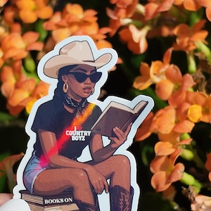 May include: A sticker featuring a woman in a cowboy hat and sunglasses reading a book. She is wearing a black t-shirt with the text "COUNTRY BOYZ" and sitting on a stack of books that read "BOOKS ON THE GROUND".