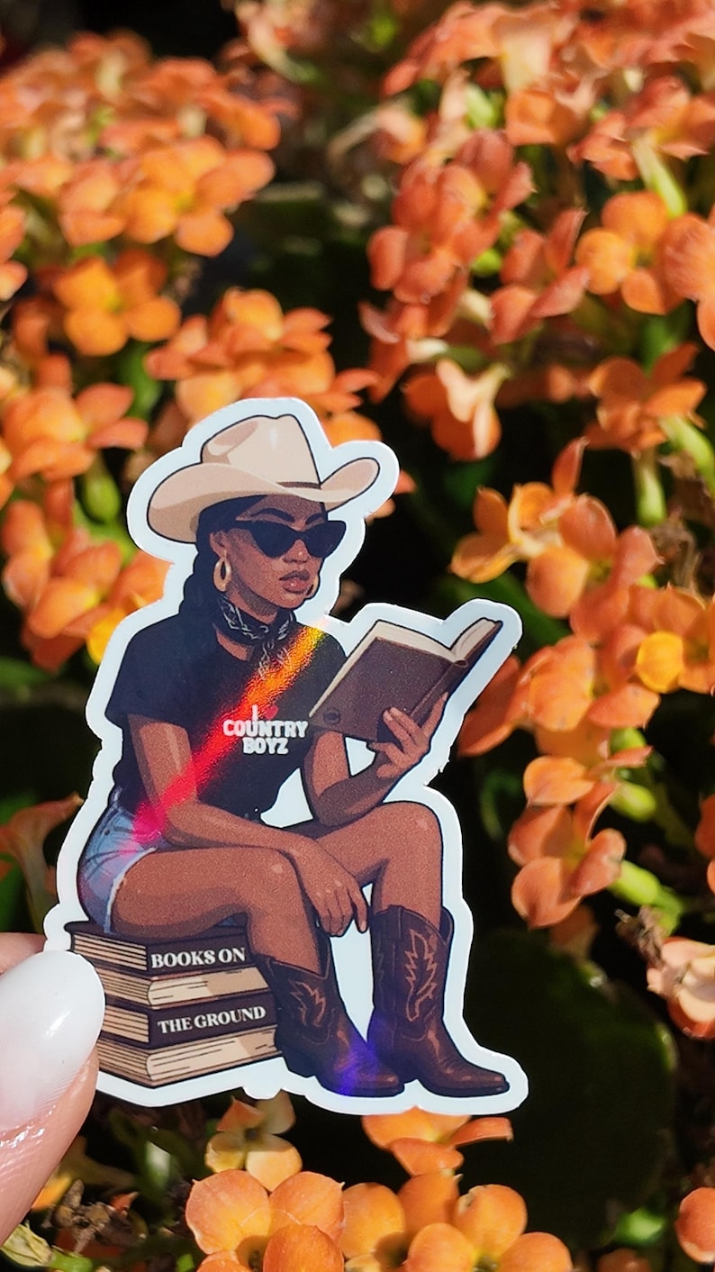 May include: A sticker featuring a woman in a cowboy hat and sunglasses reading a book. She is wearing a black t-shirt with the text "COUNTRY BOYZ" and sitting on a stack of books that read "BOOKS ON THE GROUND".