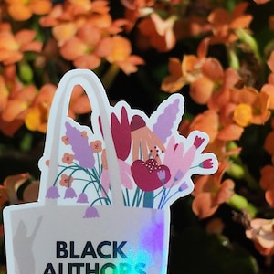 Puede incluir: Una pegatina con un bolso blanco lleno de flores coloridas. El bolso tiene el texto "BLACK AUTHORS ARE DOPE!" en letras negras e iridiscentes. El fondo son flores naranjas borrosas.