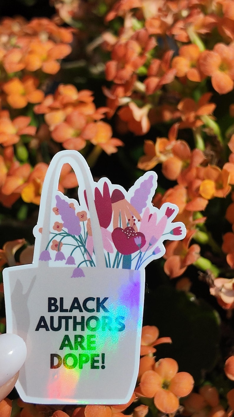 Puede incluir: Una pegatina con un bolso blanco lleno de flores coloridas. El bolso tiene el texto "BLACK AUTHORS ARE DOPE!" en letras negras e iridiscentes. El fondo son flores naranjas borrosas.