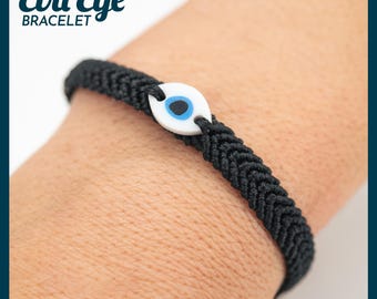 May include: Black woven bracelet featuring an evil eye charm. The charm is white with a blue eye design and a black pupil. The bracelet is worn on a wrist. The text "Evil Eye Bracelet" is in the upper left corner.