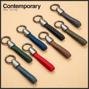 Könnte beinhalten: Eine Sammlung von Schlüsselanhängern in verschiedenen Farben, darunter Blau, Grün, Rot und Braun. Jeder Riemen hat ein gewebtes Design und einen silberfarbenen Metallring und eine Kappe. Der Text "Contemporary Key Strap" befindet sich oben links.
