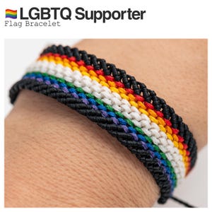 Może przedstawiać: Czarna pleciona bransoletka z tęczowymi paskami reprezentującymi flagę dumy LGBTQ+. Bransoletka jest noszona na nadgarstku osoby.  Bransoletka z flagą LGBTQ Supporter.