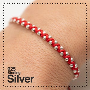 May include: A red and silver bracelet. The bracelet features a woven red cord design with evenly spaced silver beads. The text "925 Sterling Silver" is visible in the lower left corner. This bracelet is a piece of jewelry.