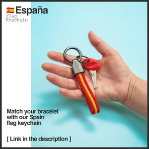 May include: A red, yellow, and black Spain flag keychain with a silver key ring and a red key. The keychain has the text "Espa&ntilde;a" and "Flag Keychain". The image also includes the text "Match your bracelet with our Spain flag keychain".