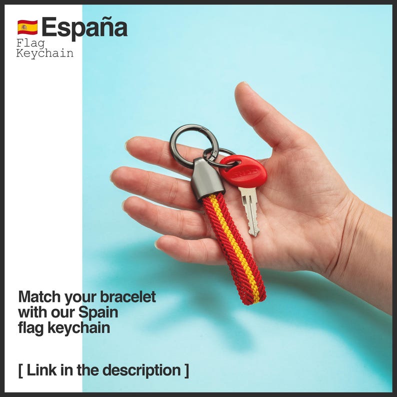May include: A red, yellow, and black Spain flag keychain with a silver key ring and a red key. The keychain has the text "Espa&ntilde;a" and "Flag Keychain". The image also includes the text "Match your bracelet with our Spain flag keychain".