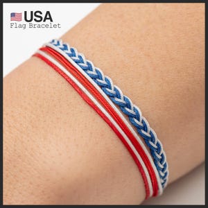 Puede incluir: Una pulsera patriótica con cordones trenzados y lisos rojos, blancos y azules. La pulsera tiene una sección trenzada azul y blanca, con cordones rojos y blancos. El texto "USA Flag Bracelet" está en la esquina superior izquierda.