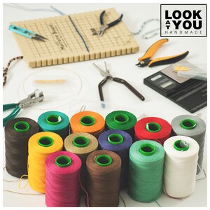 May include: A collection of colorful spools of thread, with various craft tools and a macrame board. The spools are in shades of brown, teal, orange, green, red, purple, yellow, pink, and white. Tools include pliers, scissors, and a small scale.