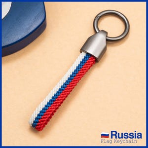 Puede incluir: Un llavero con una correa tejida con los colores de la bandera rusa: rojo, blanco y azul. El llavero tiene un anillo de metal plateado y una tapa de metal. El texto "Russia Flag Keychain" es visible.