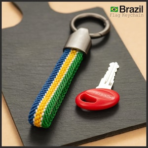 Puede incluir: Un llavero con la bandera de Brasil con una correa trenzada azul, amarilla y verde y un cierre de metal plateado. Una llave roja con "SMHS" está al lado del llavero. El texto "Brazil Flag Keychain" está en la esquina superior derecha.