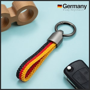 Puede incluir: Un llavero con una correa trenzada con los colores de la bandera alemana: negro, rojo y amarillo. El llavero tiene un anillo y un cierre de metal. El texto "Germany Flag Keychain" es visible.