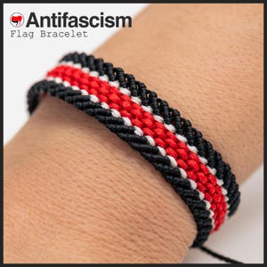 Op de afbeelding: Een gevlochten armband met een patroon van zwarte, rode en witte strepen. De armband zit om een pols. De tekst "Antifascism Flag Bracelet" is linksboven zichtbaar.