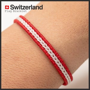 Könnte beinhalten: Ein rot-weiß geflochtenes Armband in den Farben der Schweizer Flagge. Das Armband hat einen zentralen weißen Zopf, der von roten Bändern umrandet ist. Der Text "Switzerland Flag Bracelet" ist oben links sichtbar.