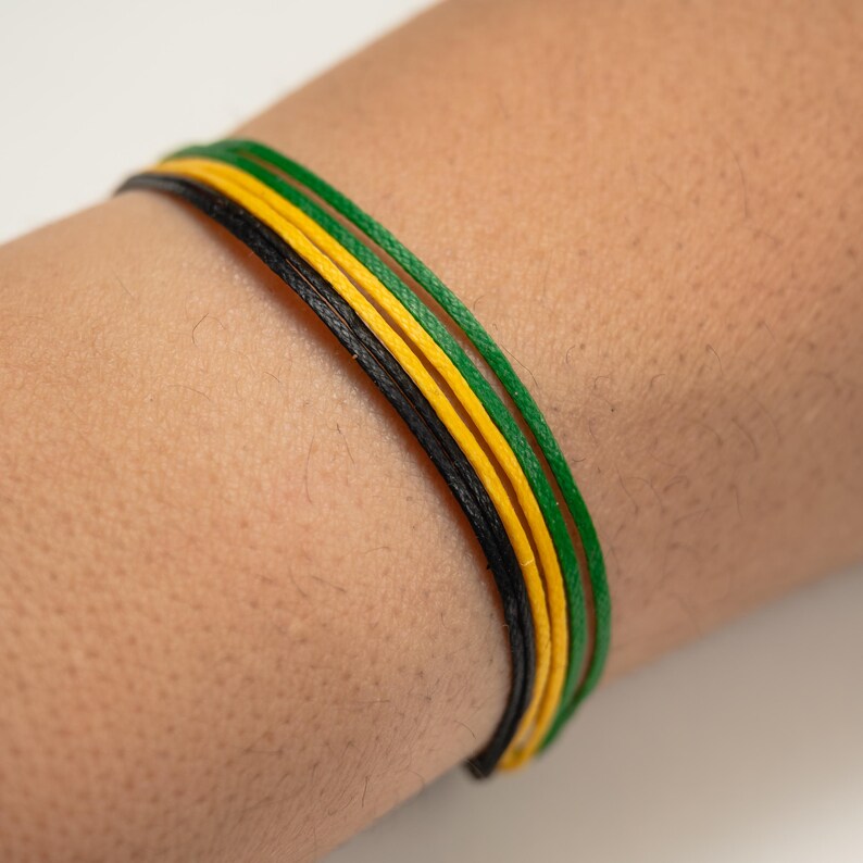 K&ouml;nnte beinhalten: Nahaufnahme eines Handgelenks mit einem Armband aus vier Schn&uuml;ren. Die Schn&uuml;re sind schwarz, gelb und gr&uuml;n, nebeneinander angeordnet. Das Armband besteht aus d&uuml;nnem, gewebtem Material.