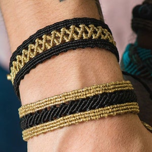 Puede incluir: Dos pulseras de macramé. La pulsera superior es negra con un diseño tejido de color dorado. La pulsera inferior es negra con detalles tejidos de color dorado. Las pulseras se llevan en la muñeca.