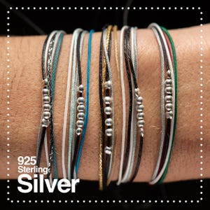 Könnte beinhalten: Eine Sammlung von mehrsträngigen Armbändern mit verschiedenfarbigen Kordeln und kleinen Silberperlen. Die Armbänder sind in den Farbtönen Schwarz, Weiß, Silber, Gold, Türkis und Grün gehalten. Der Text "925 Sterling Silver" ist unten links zu sehen.