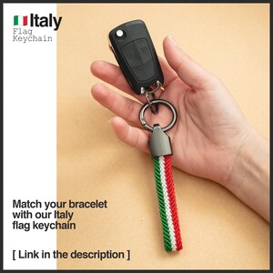 Puede incluir: Una llave de coche negra con un llavero de metal y un cord&oacute;n con los colores de la bandera italiana: verde, blanco y rojo. El llavero tiene el texto "Italy Flag Keychain". La imagen tambi&eacute;n incluye el texto "Match your bracelet with our Italy flag keychain".