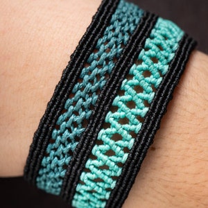 Peut inclure: Bracelet en macramé avec un motif tissé en noir et bleu sarcelle. Le bracelet a une large bande avec des motifs de nœuds complexes, créant un aspect texturé. La couleur sarcelle passe d'une nuance plus foncée à une plus claire.