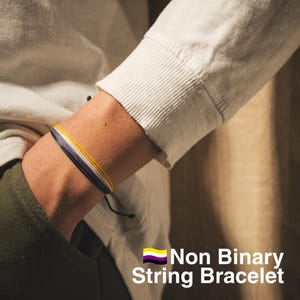 May include: A Non Binary string bracelet featuring yellow, white, purple, and black strands. The bracelet is knotted and worn on a wrist. The image includes the text "Non Binary String Bracelet".
