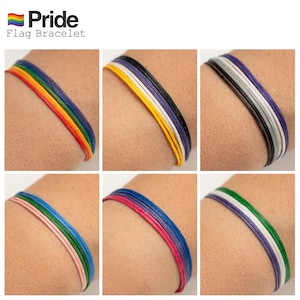 Könnte beinhalten: Ein Set aus sechs bunten Armbändern, die jeweils eine andere Pride-Flagge repräsentieren. Die Armbänder bestehen aus geflochtenen Schnüren und werden um das Handgelenk einer Person gebunden. Die dargestellten Flaggen sind die Regenbogen-Pride-Flagge, die bisexuelle Pride-Flagge, die Transgender-Pride-Flagge, die lesbische Pride-Flagge, die pansexuelle Pride-Flagge und die asexuelle Pride-Flagge.