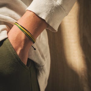 K&ouml;nnte beinhalten: Ein mehrstr&auml;ngiges Armband mit gr&uuml;nen, gelben und schwarzen Kordeln. Das Armband wird am Handgelenk getragen, die Hand in einer Tasche. Die Person tr&auml;gt ein wei&szlig;es Langarmshirt und olivgr&uuml;ne Hose.