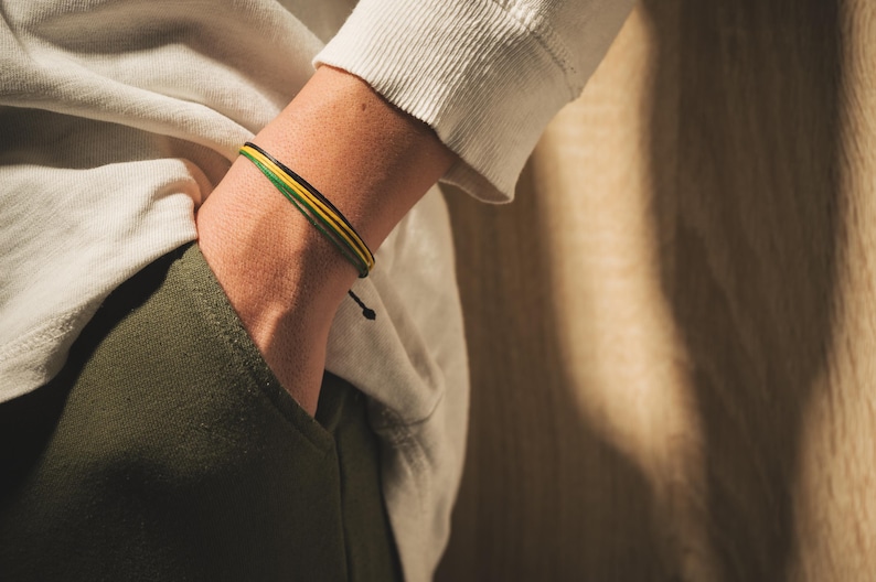 K&ouml;nnte beinhalten: Ein mehrstr&auml;ngiges Armband mit gr&uuml;nen, gelben und schwarzen Kordeln. Das Armband wird am Handgelenk getragen, die Hand in einer Tasche. Die Person tr&auml;gt ein wei&szlig;es Langarmshirt und olivgr&uuml;ne Hose.