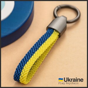 Può includere: Un portachiavi con un cinturino intrecciato nei colori della bandiera ucraina: blu e giallo. Il portachiavi ha un moschettone in metallo e una parte superiore in metallo grigio scuro. Il testo "Ukraine Flag Keychain" è visibile in basso a destra.