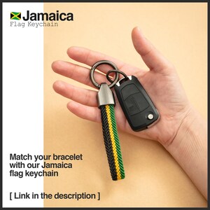 K&ouml;nnte beinhalten: Ein Jamaica-Flaggen-Schl&uuml;sselanh&auml;nger mit einem geflochtenen Band in Schwarz, Gelb und Gr&uuml;n. Der Schl&uuml;sselanh&auml;nger ist an einem silberfarbenen Metallring und einem schwarzen Autoschl&uuml;sselanh&auml;nger befestigt. Der Text "Jamaica Flag Keychain" ist sichtbar.