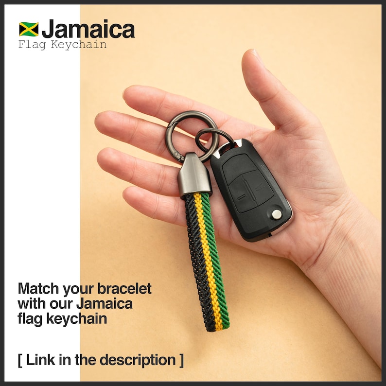 K&ouml;nnte beinhalten: Ein Jamaica-Flaggen-Schl&uuml;sselanh&auml;nger mit einem geflochtenen Band in Schwarz, Gelb und Gr&uuml;n. Der Schl&uuml;sselanh&auml;nger ist an einem silberfarbenen Metallring und einem schwarzen Autoschl&uuml;sselanh&auml;nger befestigt. Der Text "Jamaica Flag Keychain" ist sichtbar.
