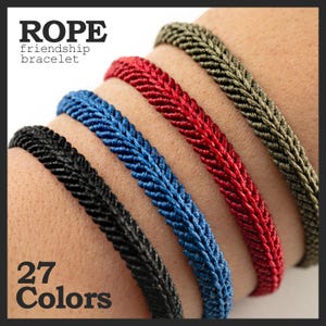 May include: Close-up of several rope friendship bracelets in black, blue, red, and olive green. The bracelets are braided and displayed on a wrist. The image includes the text "ROPE friendship bracelet" and "27 Colors".