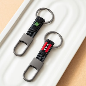 May include: Two keychains with black woven straps and metal clasps. One keychain features a green cannabis leaf on a black background. The other has a red, black, and white design. Both have silver key rings.