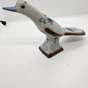 Może przedstawiać: Ceramiczna figurka ptaka z białym korpusem, brązowymi akcentami i niebieskimi kwiatowymi detalami. Ptak ma długą szyję i niebieski dziób. Podstawa jest kwadratowa z brązową obwódką i niebieskimi wzorami liści.