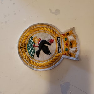 May include: Embroidered patch with a gold border and the word "Cornwallis". The patch features a black bird holding a red leaf, a green and gold design, and a castle-like structure.