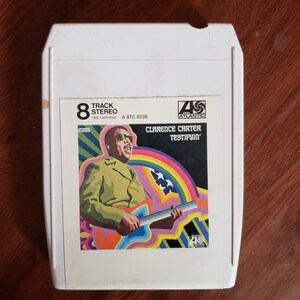 Puede incluir: Un cartucho de cinta estéreo de 8 pistas con el álbum "Testifyin'" de Clarence Carter. La carátula del álbum muestra una colorida ilustración de Carter tocando la guitarra, con un arcoíris y estrellas de fondo. El cartucho es blanco con el logo de Atlantic Records.