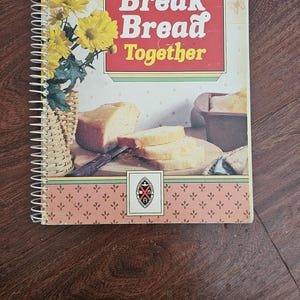 Può includere: Un libro di cucina rilegato a spirale intitolato "Let's Break Bread Together" con una copertina rossa e gialla. La copertina presenta illustrazioni di pane, fiori e un cestino. Il libro è di The United Churches in Canada.