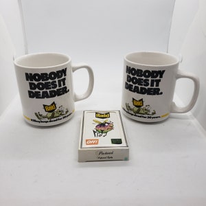Puede incluir: Dos tazas de cerámica blancas con el texto "NOBODY DOES IT DEADER" y el logotipo de Raid. Un mazo de naipes "Off!" de plasticard está delante de las tazas. Las tazas y las cartas están sobre una superficie blanca.
