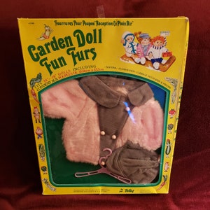 Puede incluir: Un conjunto de ropa de muñeca vintage en una caja amarilla. El conjunto incluye una chaqueta de piel sintética rosa, un cuello y una falda de pana gris y un sombrero a juego. La caja dice "Garden Doll Fun Furs" y "Para muñecas de 35,56cm - 45,7cm".