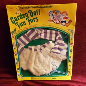 Puede incluir: Una caja amarilla vintage con el texto "Garden Doll Fun Furs" e ilustraciones. En el interior, un conjunto de piel sintética blanca con un gorro y bufanda a juego a rayas moradas y blancas, diseñado para muñecas de 35,56 cm a 45,7 cm.