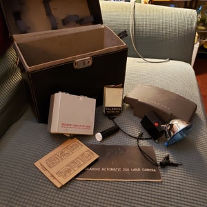 Peut inclure: Un kit d'appareil photo Polaroid vintage, comprenant un étui en similicuir noir, un kit de gros plan blanc et divers accessoires. Le kit comprend un flash, des filtres et des manuels d'instructions. Le texte "Polaroid Automatic 250 Land Camera" est visible.