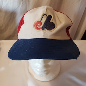 May include: A vintage baseball cap with a white front panel featuring a red, blue, and white embroidered logo. The cap has a navy blue peak and a red mesh back. The hat is displayed on a white mannequin head.