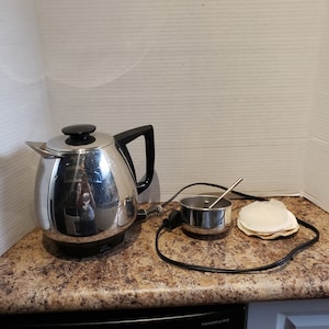 Puede incluir: Una cafetera eléctrica de acero inoxidable con asa y tapa negras. Un pequeño recipiente de acero inoxidable con una cuchara y una pila de filtros de café están junto a la cafetera. Los artículos están sobre una encimera.
