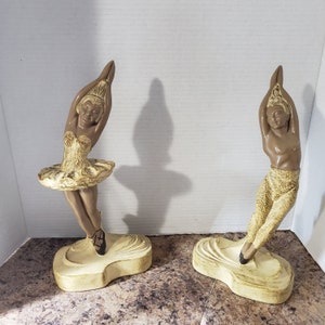 Puede incluir: Dos figuras de bailarines de ballet de cerámica. Una bailarina lleva un tutú dorado y la otra pantalones dorados. Ambas figuras están en una pose elegante con los brazos levantados. Las figuras son de color beige y marrón, con detalles negros en los zapatos. Se apoyan en bases color crema.