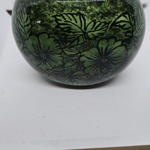 Könnte beinhalten: Ein runder, grüner Keramik-Blumentopf mit floralem Design. Der Topf hat einen dunkelgrünen Hintergrund mit schwarzen Blumen- und Blätternkonturen. Der Innenraum ist hellbeige.