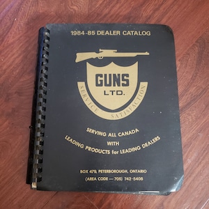 Op de afbeelding: Een zwarte 1984-85 Dealer Catalogus met een spiraalbinding. De omslag heeft een gouden embleem met een geweersilhouet en de tekst "GUNS LTD." en "SERVICE SATISFACTION". Extra tekst bevat "SERVING ALL CANADA" en contactgegevens.